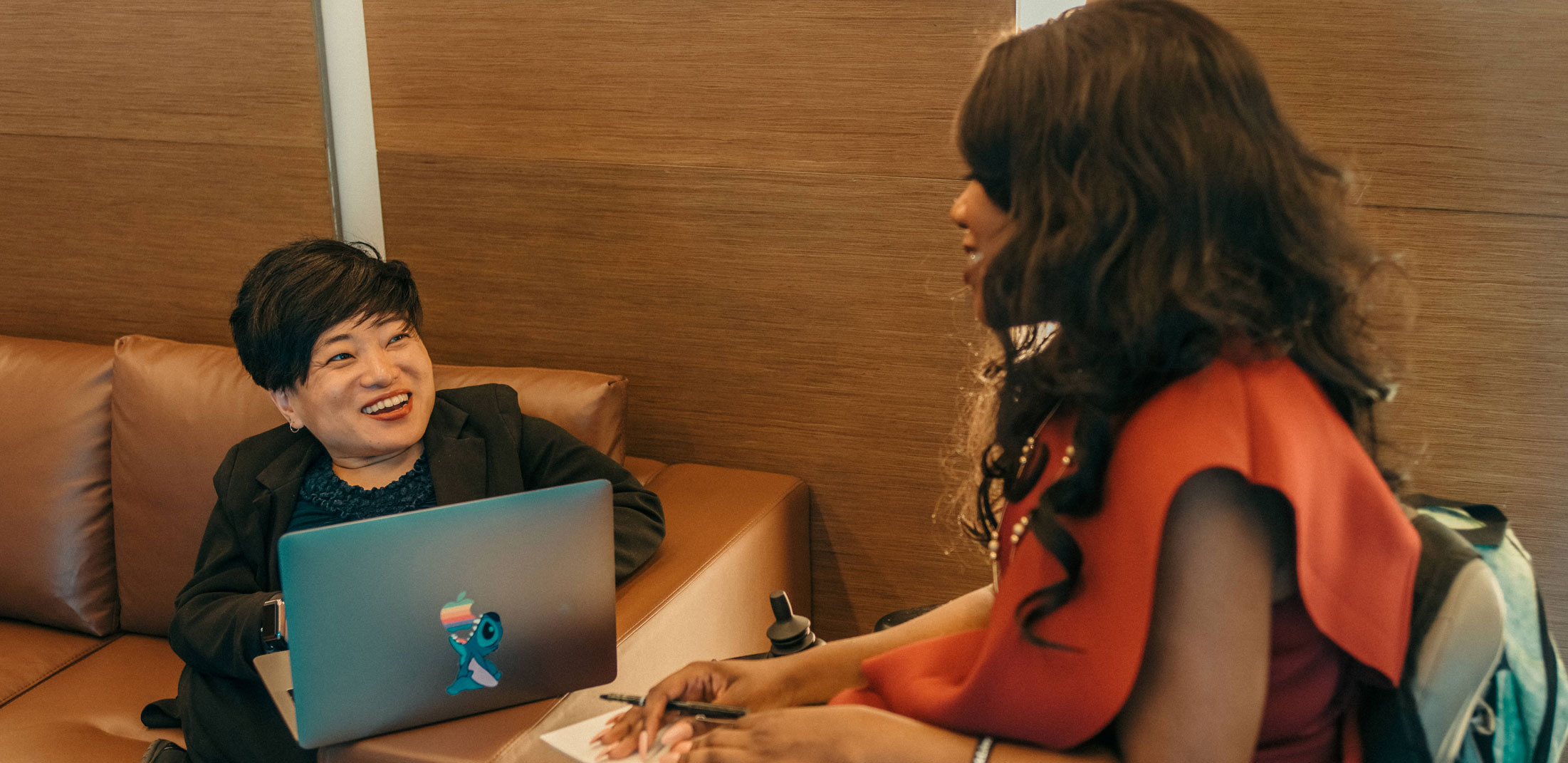 An asian woman sitting on her laptop looking towards a black woman on her right. They are smiling and engaged in conversation.